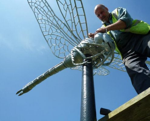 Gary stand on a platform bolting on giant dragonfly wings a giant dragonfly body which is mounted on a tall metal post, at Hounslow Heath parklands.. Public Art London